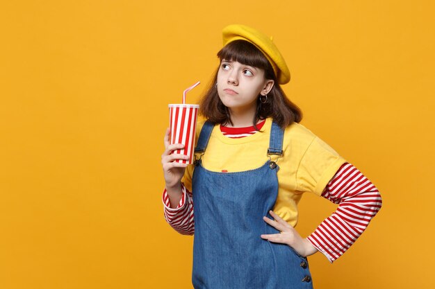 Desagradou a adolescente no vestido de denim de boina francesa segurando o copo plástico de cola ou refrigerante isolado no fundo da parede amarela no estúdio. Conceito de estilo de vida de emoções sinceras de pessoas. Mock up espaço de cópia.