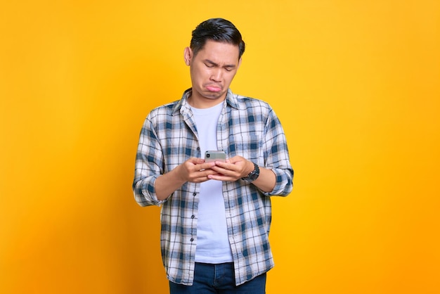 Desapontado jovem asiático em camisa xadrez usando telefone celular lendo más notícias isoladas em fundo amarelo