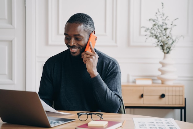 Un desarrollador afroamericano positivo y alegre sentado en el escritorio mirando la pantalla de la computadora hablando en un teléfono inteligente con el cliente para discutir la aplicación que trabaja desde casa para resolver problemas comerciales a distancia
