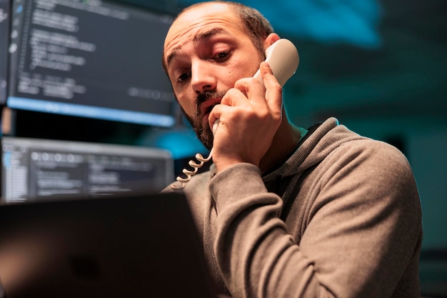 Desarrollador de aplicaciones hablando por teléfono fijo después de horas, contestando el teléfono del cable de la oficina para programar el código en la ventana de la terminal. Administrador de la base de datos usando la línea telefónica para desarrollar una nueva interfaz.