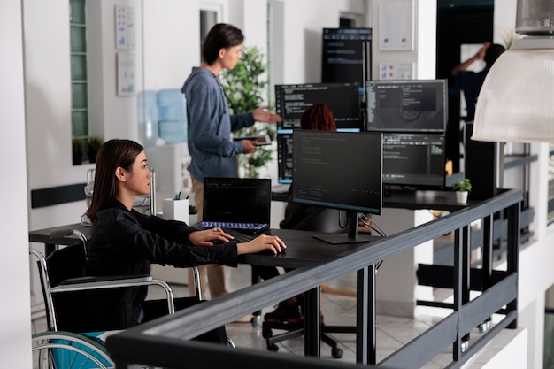 Foto desarrollador de software enfocado en silla de ruedas escribiendo código de servidor en la sala de la agencia de codificación, utilizando el sistema de seguridad. ingeniero asiático con interfaz de usuario de programación de discapacidad física ai office.