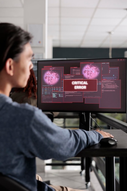 Desarrollador de software que ha pirateado el servidor web mirando la computadora con un error crítico en la pantalla en la oficina de programación. Ingeniero de sistemas asiático tratando de resolver problemas de piratería, seguridad de cortafuegos.