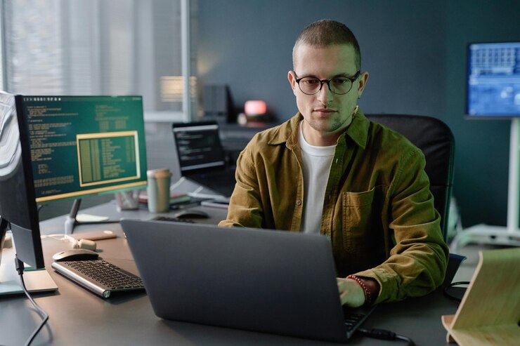 Foto desarrollador de ti masculino con gafas codificando en la computadora portátil mientras está sentado en el escritorio de la oficina
