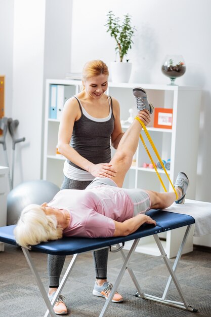 Desarrollar los músculos de las piernas. Mujer amigable positiva de pie cerca de su paciente mientras lo ayuda a desarrollar los músculos de sus piernas