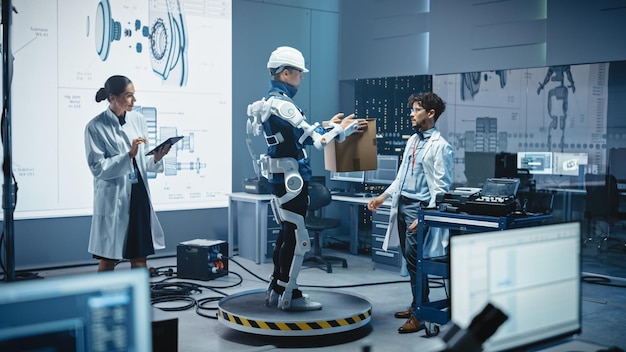 En el desarrollo, los ingenieros de laboratorio trabajan en un exoesqueleto robótico con una persona que lo prueba sosteniendo una pesada caja de cartón diseñando un exoesuit portátil para ayudar a los trabajadores de almacén de trabajo duro