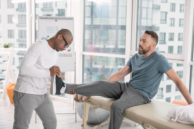 Desarrollo muscular. Bonito hombre brutal sentado levantando la pierna mientras desarrolla sus músculos