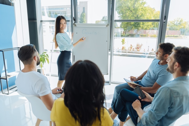 Desarrollo profesional. Atractiva mujer joven positiva de pie cerca de la pizarra y mirando a su grupo mientras les presenta información