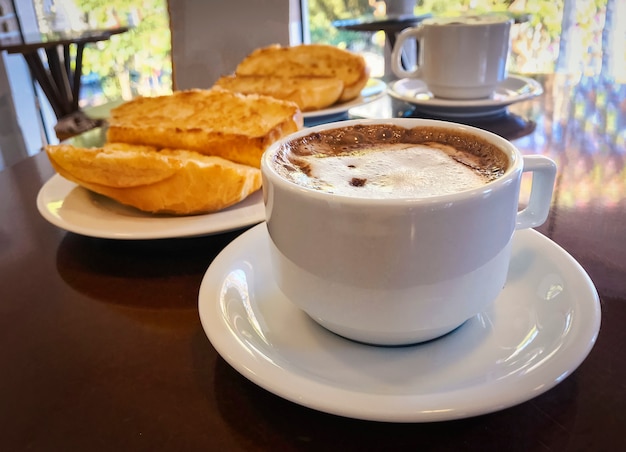 Desayune en el Brasil con el pan francés tostado con mantequilla en la placa con capuccino en la tabla.