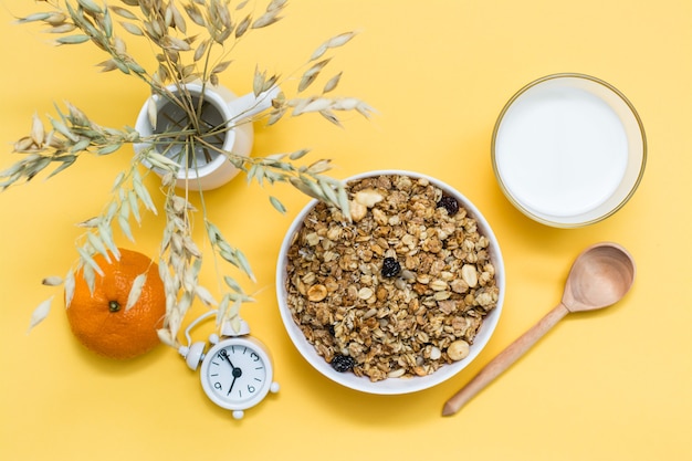 Desayuno abundante y saludable. Granola al horno en un tazón, vaso de leche, naranja, cuchara de madera y despertador sobre superficie amarilla. Vista superior