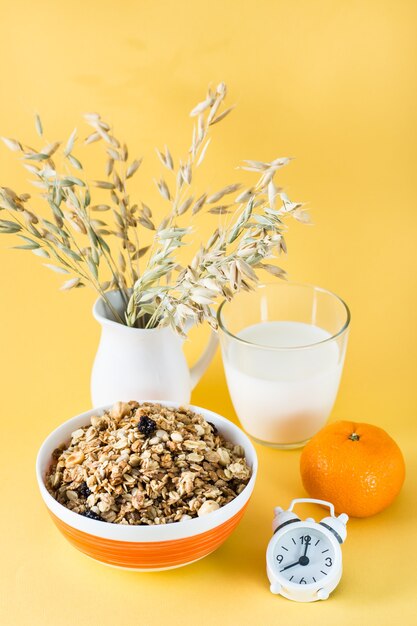 Desayuno abundante y saludable. Granola al horno en un tazón, vaso de leche, naranja y despertador sobre superficie amarilla. Vista vertical