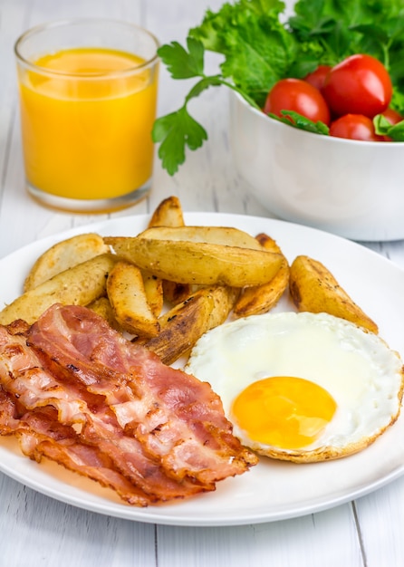 Desayuno abundante con tocino, huevo frito, papa y un vaso de jugo de naranja.