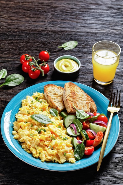 Desayuno americano de huevos revueltos con pepino, ensalada de tomate y espinacas, pan tostado y un vaso de jugo de naranja fresco en un plato azul sobre una mesa rústica de madera oscura, vista vertical