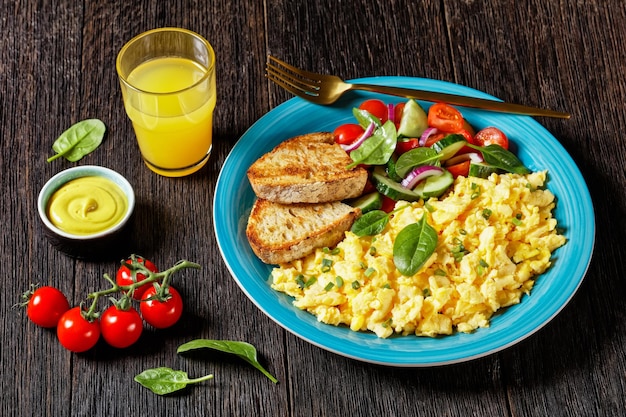 Desayuno americano de huevos revueltos con pepino, ensalada de tomate y espinacas, pan tostado y un vaso de jugo de naranja fresco en un plato azul sobre una mesa rústica de madera oscura.