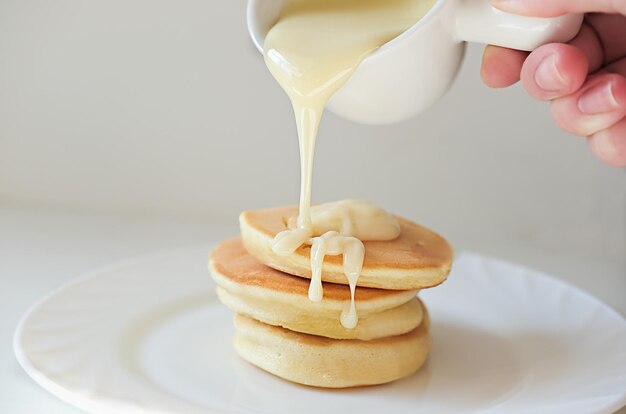 Desayuno asiático de moda, panqueques de soufflé japonés esponjosos, mano sosteniendo sauseboat vertiendo salsa de crema sobre tres pasteles calientes en un plato