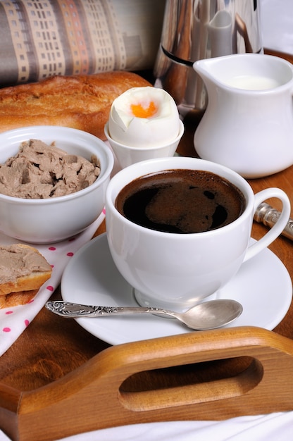 Desayuno en bandeja con una taza de café, tostadas con paté de hígado, huevo pasado por agua y un periódico