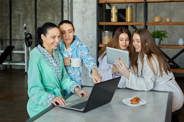 Desayuno en batas de baño, familia de cuatro mujeres charlando tomando té y viendo información en dispositivos móviles en la mesa del comedor.