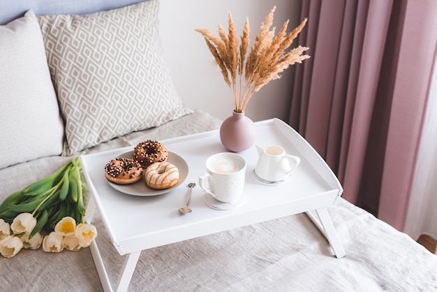 Desayuno en la cama con taza de capuchino, donas y tulipanes blancos.