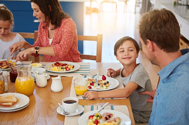 Foto desayuno de comida y una familia juguetona en el comedor de su casa juntos para la salud o la nutrición madre padre y niños hermanos lindos comiendo en una mesa en su apartamento por amor o vínculo