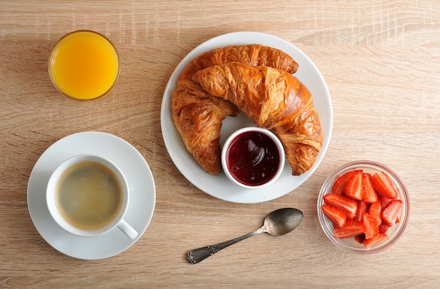 Desayuno continental con café, croissant con mermelada, fresas y jugo de naranja sobre superficie de madera.