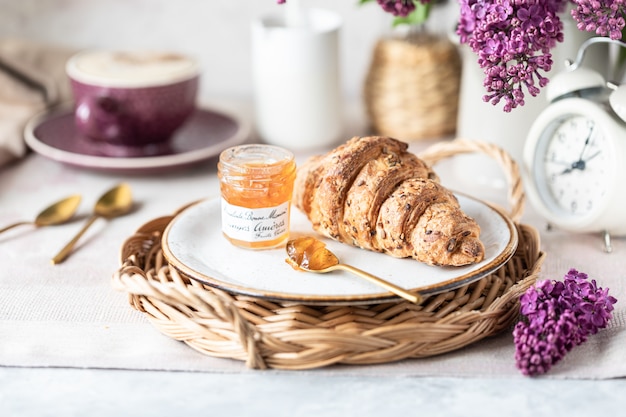 Desayuno Croissant francés, mermelada, taza de café, leche y crema y flores de color lila. Mañana