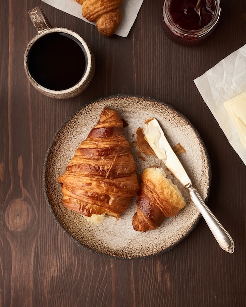 Desayuno con croissant y rebanada en el plato, taza de café, mermelada y mantequilla.