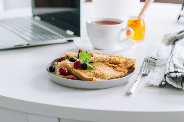 Desayuno delicioso. Mesa de escritorio blanca con laptop y crepes frescos, taza de té