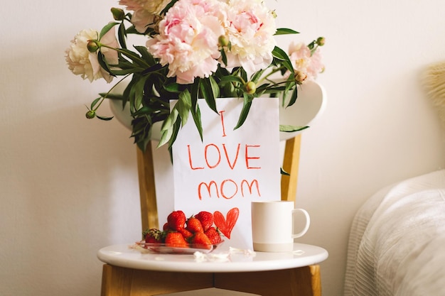 Desayuno para el Día de la Madre Plato blanco en forma de corazón con fresas frescas taza de regalo de café y ramo de peonías con regalo en la cama Composición de bodegones Feliz Día de la Madre