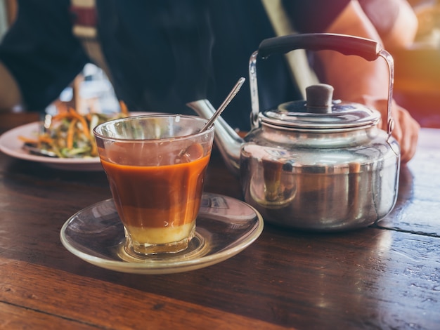 Desayuno estilo sureño tailandés, té tailandés caliente en vidrio y tetera vintage en mesa de madera.