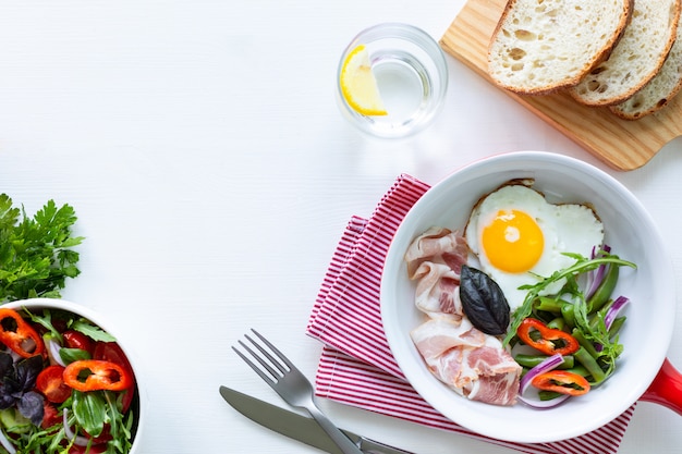Desayuno europeo: huevo en forma de corazón, tocino, judías verdes sobre una mesa blanca. Enfoque selectivo. Vista desde arriba. Copie el espacio.