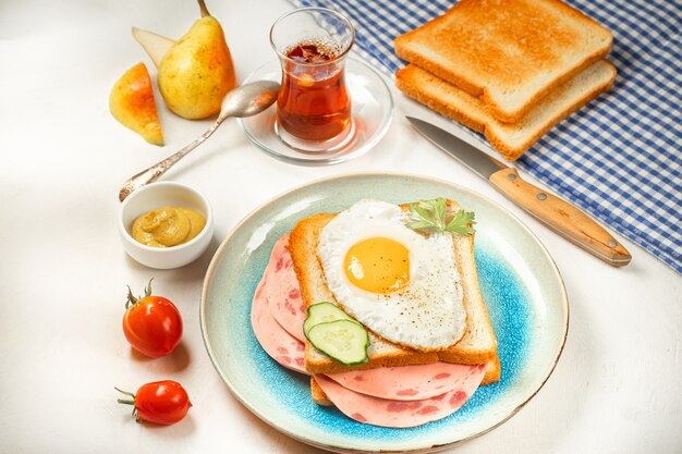 Desayuno fácil: huevo frito en sándwich con salchicha en plato con tomate, mostaza, jamón, tostadas, té de hierbas y peras. Fondo blanco. Vista superior.