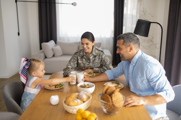 Desayuno en familia. Radiante mujer militar feliz sonriendo ampliamente mientras desayuna con la familia