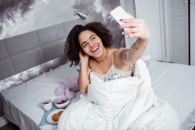 Desayuno francés cercano. Radiante chica rizada fotografiando su desayuno en la cama con el teléfono inteligente en la mano