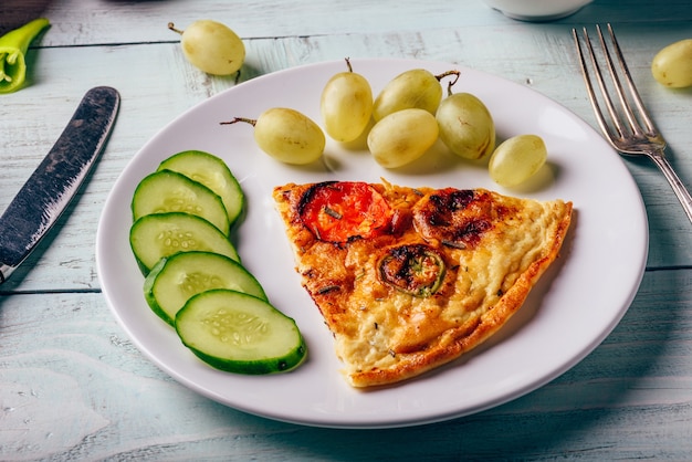 Desayuno frittata con rodajas de pepino y uvas verdes en plato blanco sobre mesa de madera.