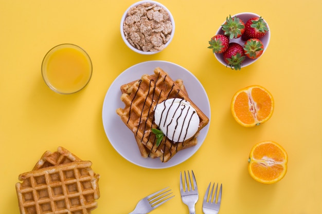 Desayuno de gofres con jugo de helado, fruta y cereales en mesa amarilla