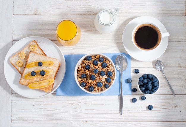 Desayuno con granola y arándanos, café con jugo de naranja y tostadas al horno sobre la mesa blanca