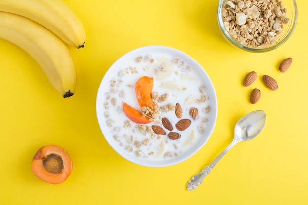 Desayuno de granola con frutas de leche y almendras en el tazón blanco sobre fondo amarillo Vista superior Primer plano