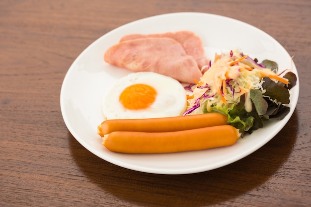 Foto desayuno con huevo, jamón, salchicha en plato blanco en la mesa de madera