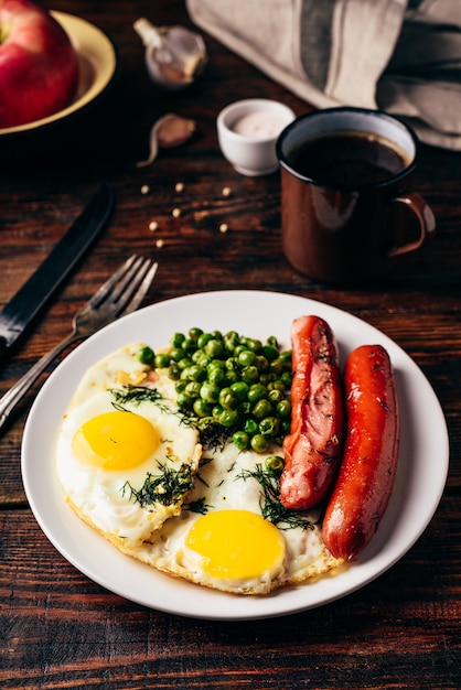 Desayuno con huevos fritos, salchichas y guisantes en la placa blanca.