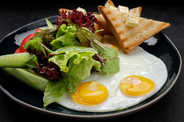 Desayuno Huevos fritos con tostadas con mantequilla y verduras pepinos y tomates En un plato oscuro sobre un fondo oscuro