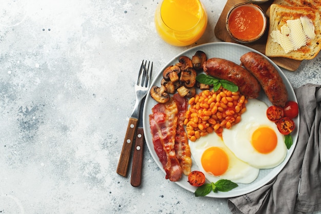 Desayuno inglés completo en un plato con huevos fritos, salchichas, tocino, frijoles, tostadas y café sobre fondo de piedra clara. Con espacio de copia. Vista superior.