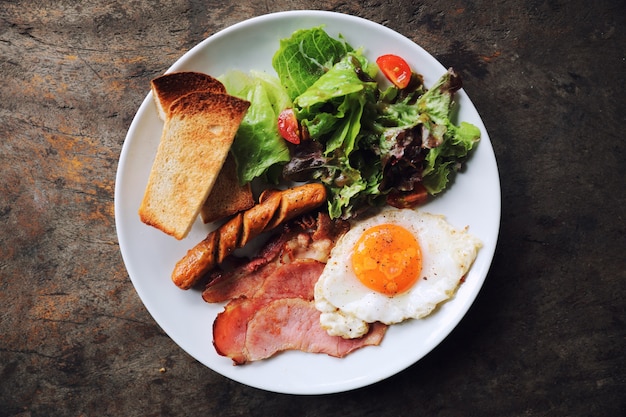 Desayuno inglés con huevo frito, jamón, salchicha, tocino, ensalada y tostadas en la placa blanca.