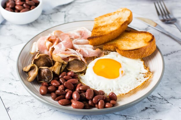 Desayuno inglés con huevo frito tocino frijoles champiñones y tostadas en un plato sobre la mesa Primer plano