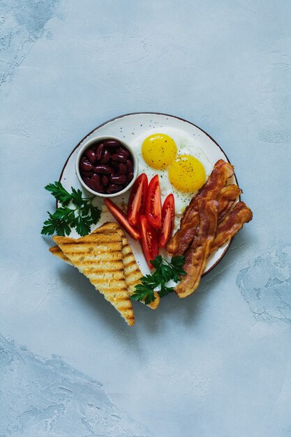 Desayuno inglés tradicional con huevos fritos, tocino, frijoles, tostadas y tomates sobre hormigón gris