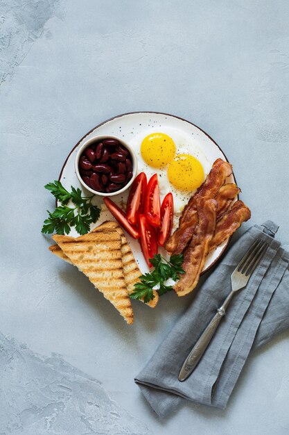 Desayuno inglés tradicional con huevos fritos, tocino, frijoles, tostadas y tomates sobre hormigón gris