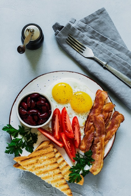 Desayuno inglés tradicional con huevos fritos, tocino, frijoles, tostadas y tomates sobre hormigón gris