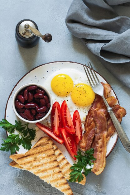 Desayuno inglés tradicional con huevos fritos, tocino, frijoles, tostadas y tomates sobre hormigón gris