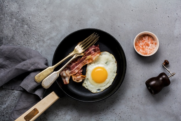 Desayuno inglés tradicional con huevos fritos y tocino en sartén de hierro fundido sobre fondo de hormigón gris antiguo. Vista superior.