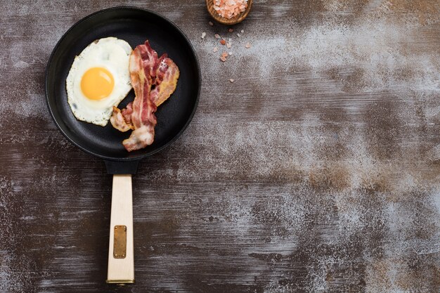 Desayuno inglés tradicional con huevos fritos y tocino en sartén de hierro fundido sobre fondo de hormigón oscuro. Vista superior.