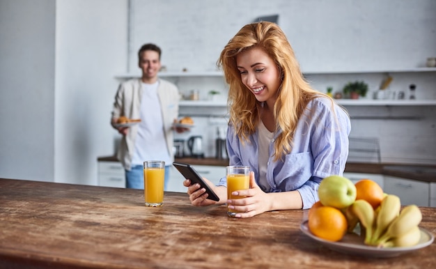 Desayuno de una joven pareja de enamorados en la cocina de casa. Hermosa chica con un teléfono en la mano bebe jugo.