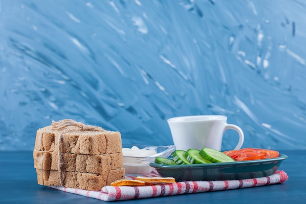 Un desayuno ligero, una taza de té, un tazón de rodajas de pepino y tomates y pan sobre un paño de cocina, sobre la mesa azul.